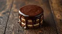 Generate an simple image of matilda micro cake cylindrical shape layered topped with chocolate ganache plated in wooden table.webp